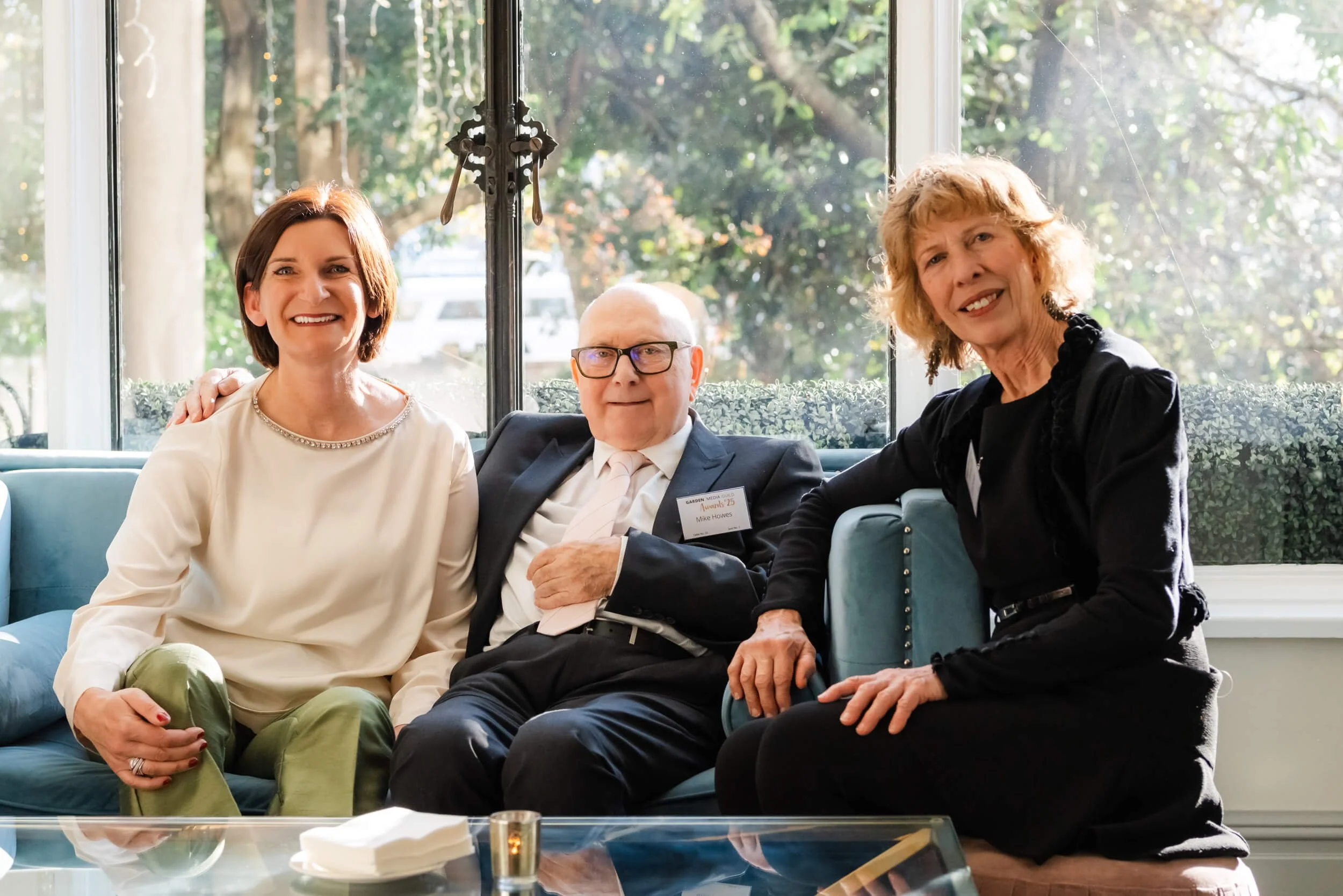 Three people sitting on a blue couch inside a house with a large window behind them. The woman on the left is wearing a cream blouse and green pants, the man in the middle is wearing glasses and a suit with a name badge, and the woman on the right is