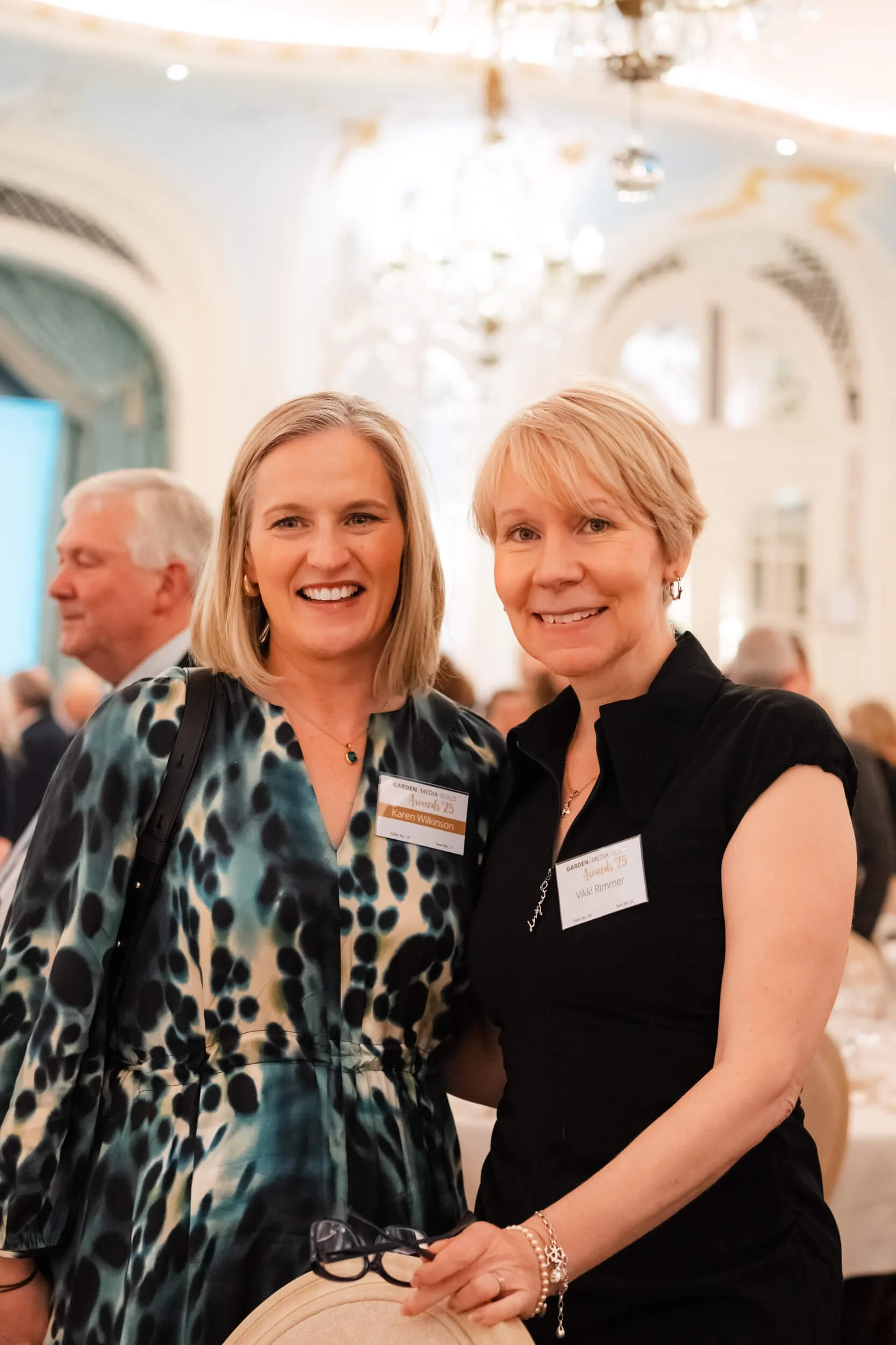 Two women smiling at a formal event, standing close together, with name tags, in a large ornate room with high ceilings and chandeliers.