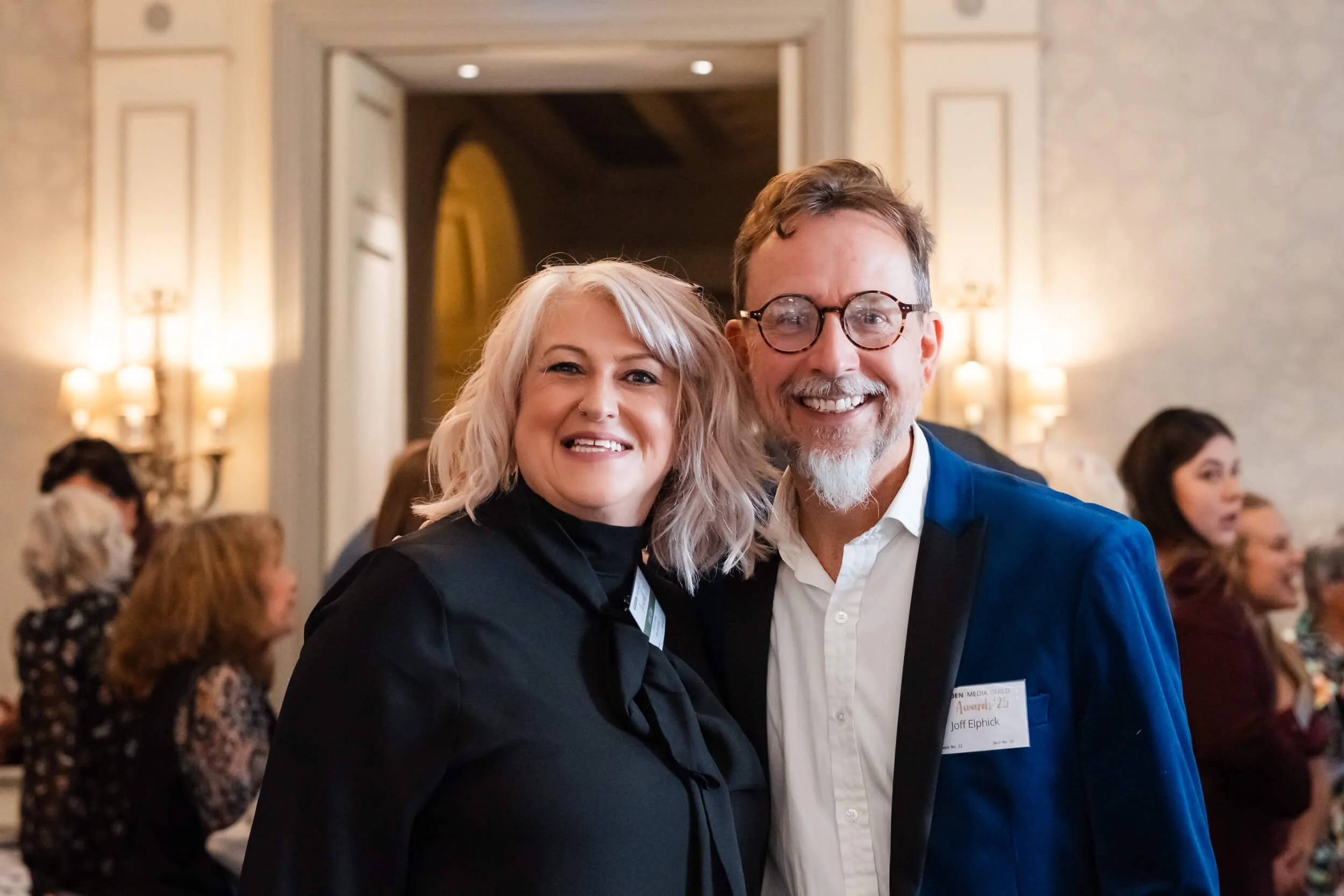 A smiling man with round glasses and a white beard wearing a blue jacket and white shirt, standing next to a smiling woman with shoulder-length blonde hair, dressed in black, at a formal event in a decorated indoor setting with other people in the ba