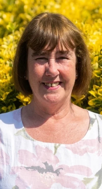 A smiling woman with short brown hair standing outdoors in front of yellow flowering bushes.