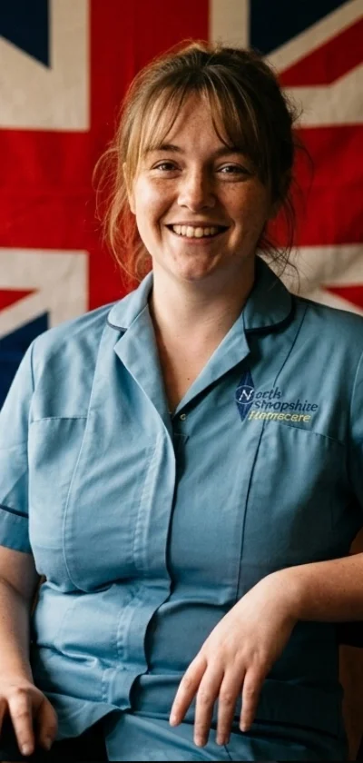 A woman wearing a light blue nurse uniform with the North Shropshire Vets and Nursecare logo, standing in front of a British flag, smiling at the camera.