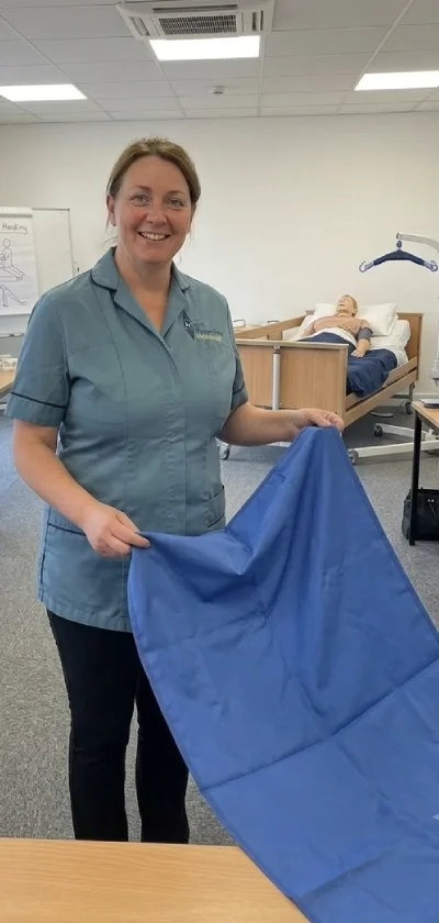 A woman nurse in a blue uniform holding a blue cloth sheet, standing in a clinical training room with a hospital bed and a medical mannequin in the background.