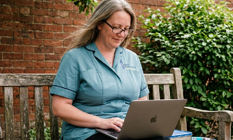 Woman in teal uniform sitting on a wooden bench outdoors, working on a silver laptop, with green bushes and a brick wall in the background.