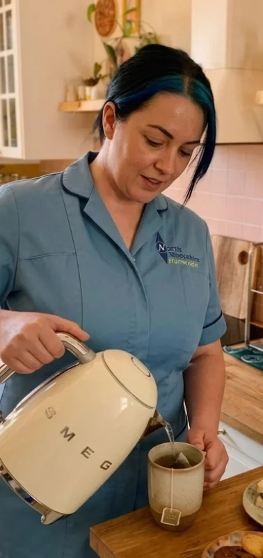 A woman with blue hair wearing a blue shirt is pouring hot water from an off-white kettle into a mug on a wooden countertop.