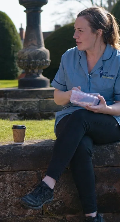 A woman sitting on a stone bench outdoors, holding a plastic container, with a disposable coffee cup on the bench beside her, and a fountain in the background.