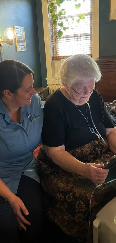 A young woman with dark hair and a blue shirt sitting beside an elderly woman with white hair, who is wearing a black top and a patterned skirt, looking at a device together in a cozy room with a window and houseplants.