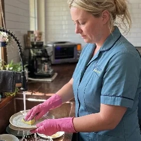 A woman washing dishes at the kitchen sink, wearing a blue shirt and pink rubber gloves.