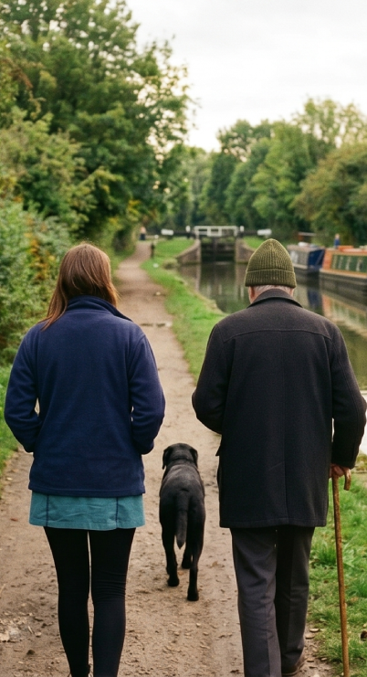 Two people walking with a dog along a canal path lined with trees and greenery.