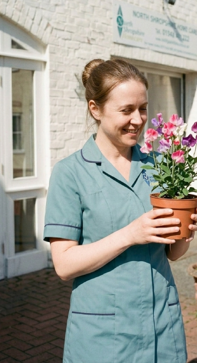A woman in a light blue nurse uniform holding a pot of pink and purple flowers outdoors.