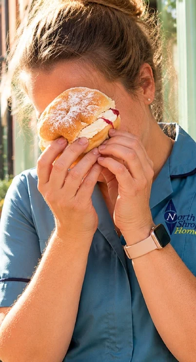 A woman holding and eating a cream-filled pastry with powdered sugar dusting, outdoors near a window.
