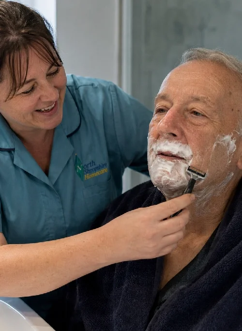 A woman in a light blue uniform is shaving an elderly man with white hair and a beard, who is sitting with a towel around his shoulders.