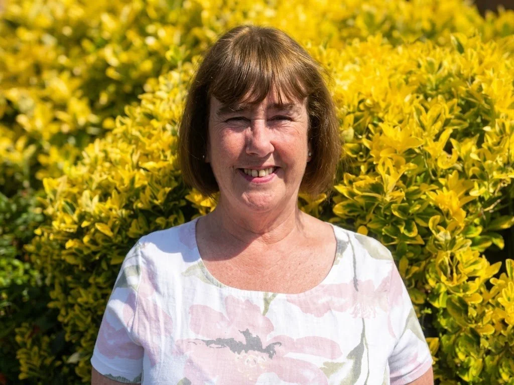 A woman with short brown hair smiling outdoors, standing in front of a lush, yellow-green shrub on a sunny day.