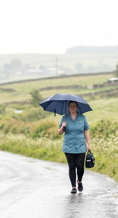 A woman walks outdoors on a rainy day, holding a blue umbrella and carrying a small bag, with a green countryside background.