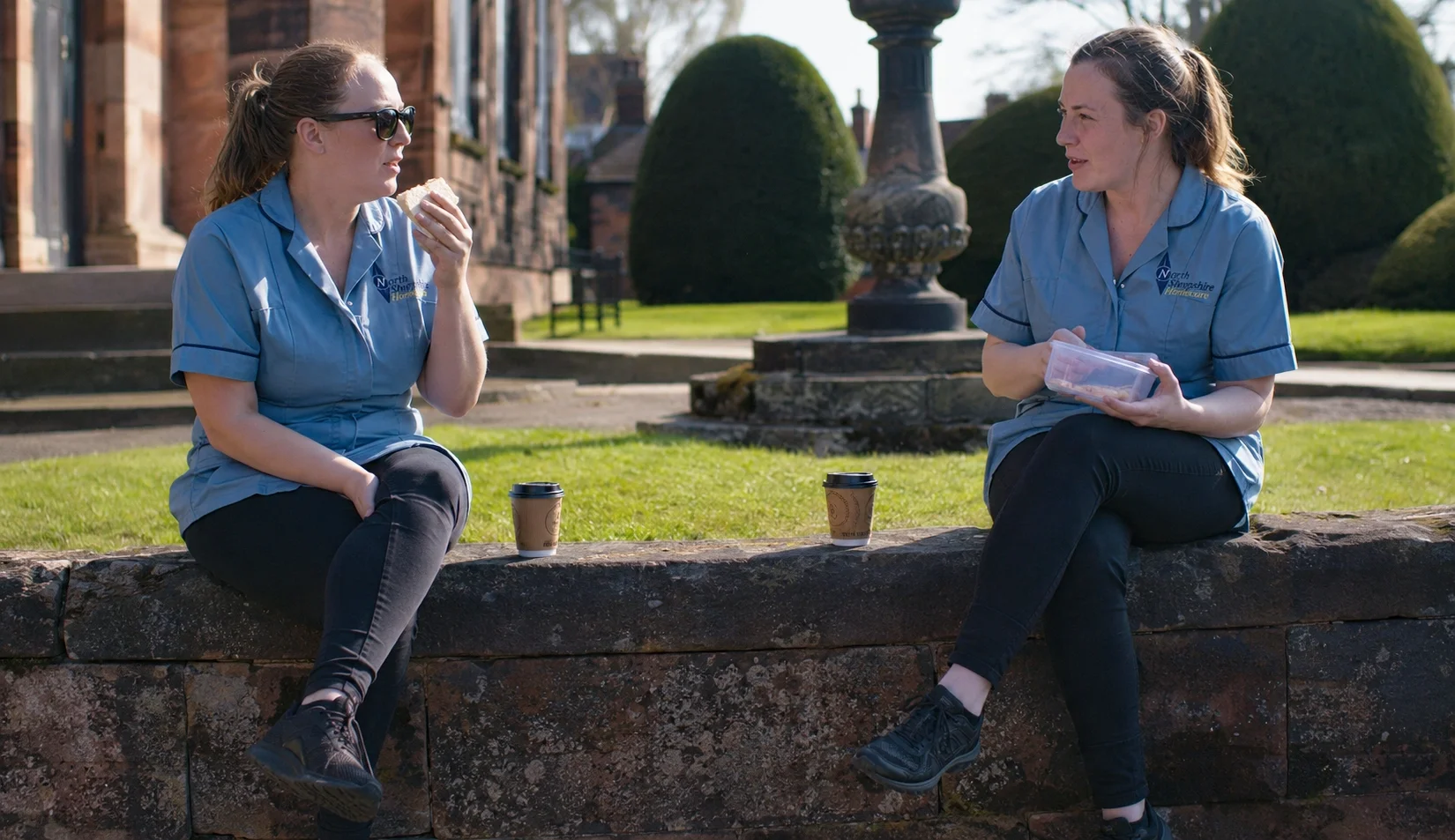 Two women in blue uniforms sit on a stone wall outdoors, having a conversation while holding food and drinks. They are in a park or garden area with manicured bushes and historic buildings in the background.