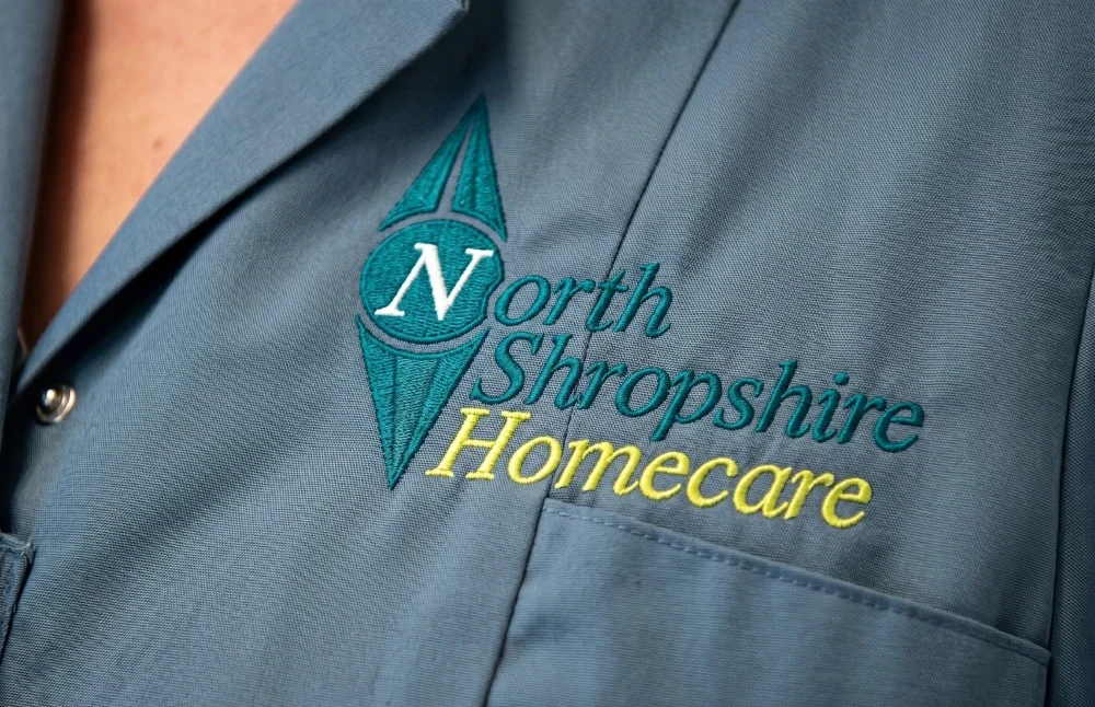 Close-up of a blue shirt with an embroidered logo reading 'North Shropshire Homecare' and a compass design.