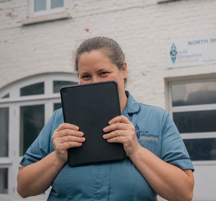 A woman smiling behind a black tablet inside a white brick building with windows, wearing a blue uniform with a North Shropshire Homecare logo.