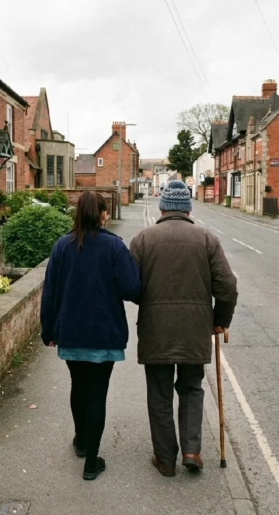 A younger woman and an elderly man walking together down a neighborhood street, holding hands. The man is using a cane and wearing a beanie. The street is lined with brick houses and greenery.