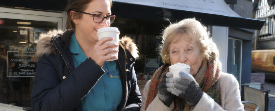 A young woman and an elderly woman drinking hot beverages outdoors on a city street.