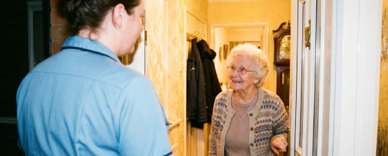 An elderly woman with white hair and glasses smiling and talking to a woman in a blue shirt at her doorway.
