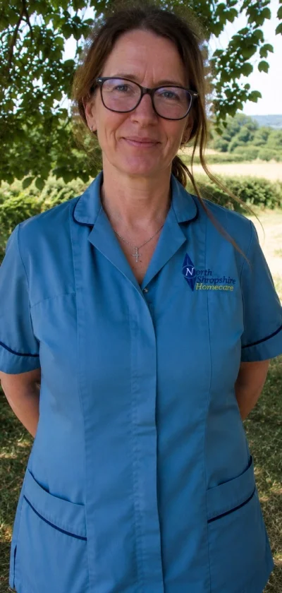 A woman with glasses in a blue uniform stands outdoors in front of green trees and a field, smiling slightly.