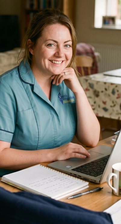 A woman in a blue uniform smiling at the camera, sitting at a table with a laptop, notebook, pen, and a mug in a cozy room.