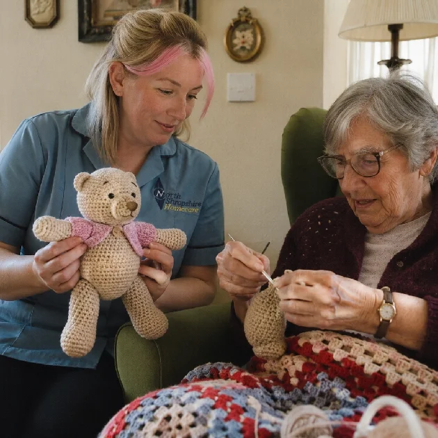 A young woman and an elderly woman are sitting together, knitting a blanket. The young woman is holding a crocheted teddy bear. They are in a cozy living room.