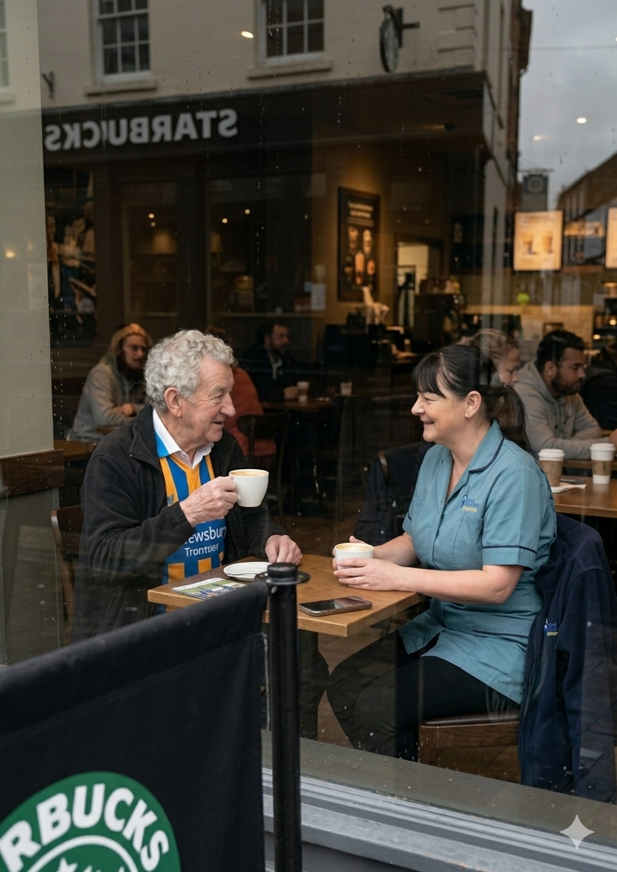 An elderly man and a woman in medical scrubs are sitting inside a coffee shop, smiling and talking while holding cups of coffee. Other customers are seated in the background.