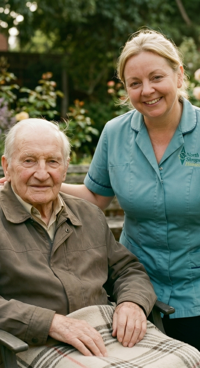 An elderly man sitting outdoors in a chair with a blanket, smiling, while a caregiver in scrubs standing next to him smiles in a garden setting with trees and flowers.