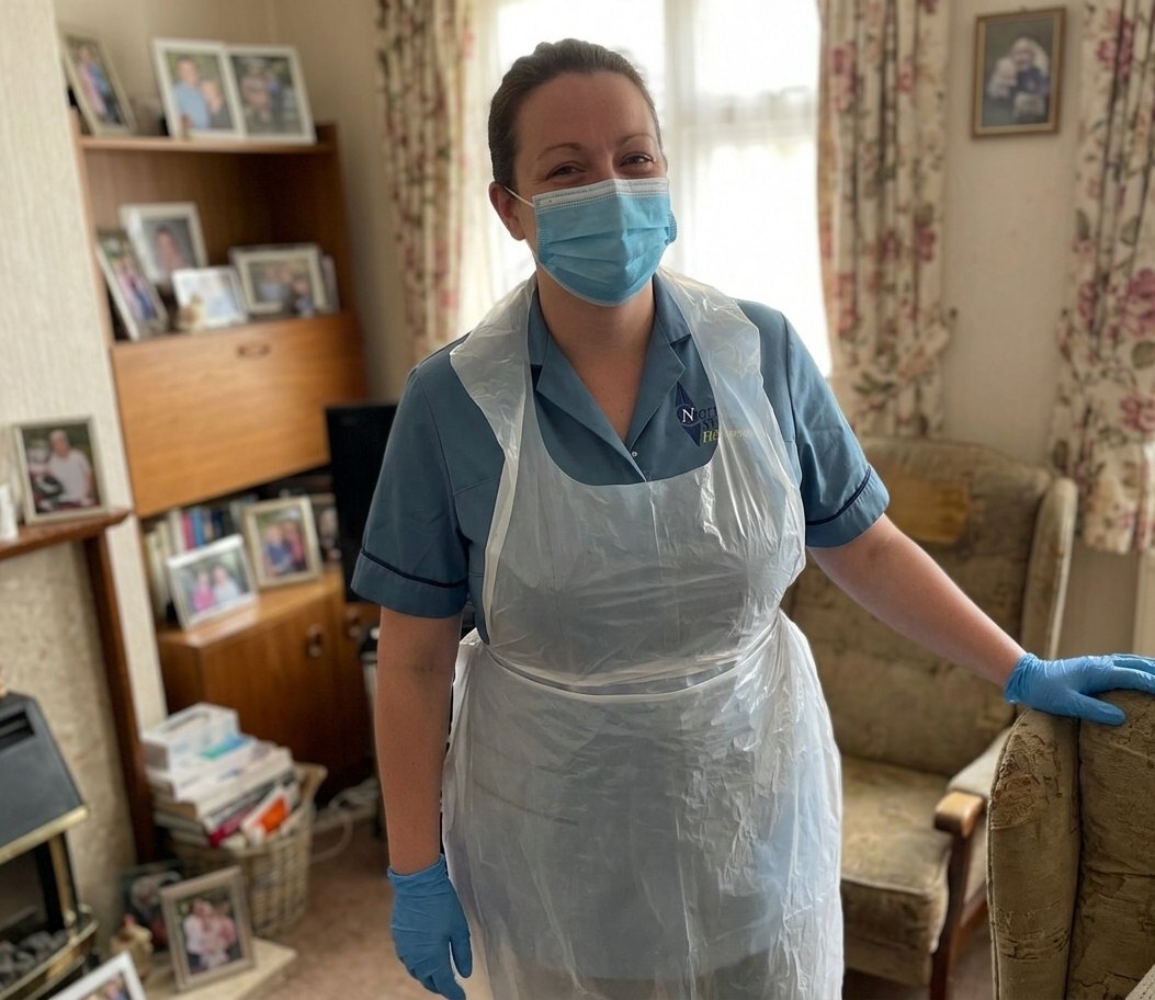 A woman in healthcare uniform, wearing a face mask, gloves, and a plastic apron, standing in a living room with floral curtains and family photos on shelves.