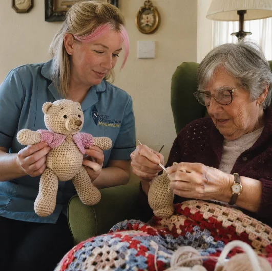 A young woman and an elderly woman are sitting together. The young woman is holding a knitted teddy bear, while the elderly woman is knitting. They are in a cozy living room with a blanket and a lamp in the background.