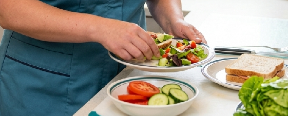 Person preparing a fresh vegetable salad with slices of tomato and cucumber, and whole grain bread on the side.