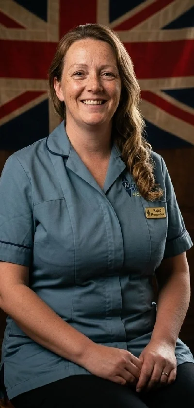 A woman in a blue uniform with a name tag seated in front of a Union Jack flag. She is smiling with long hair and appears to be in a professional setting.