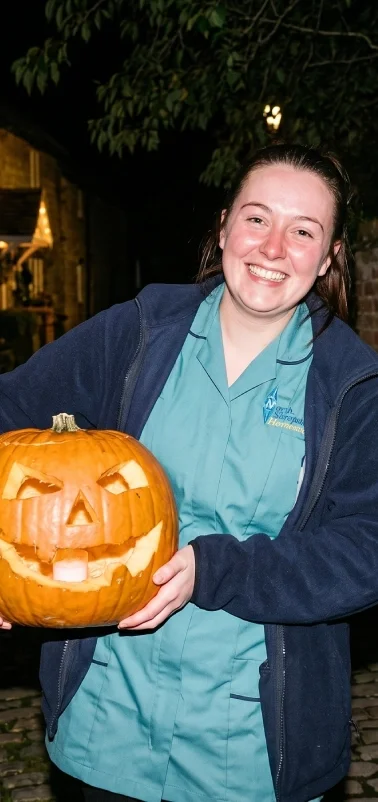 Smiling woman holding a carved pumpkin with a jack-o'-lantern face, outdoor nighttime setting.