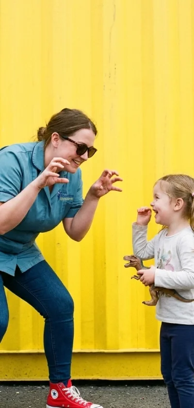 A woman wearing sunglasses and a blue shirt is playing with a young girl holding a small dinosaur toy in front of a yellow wall.