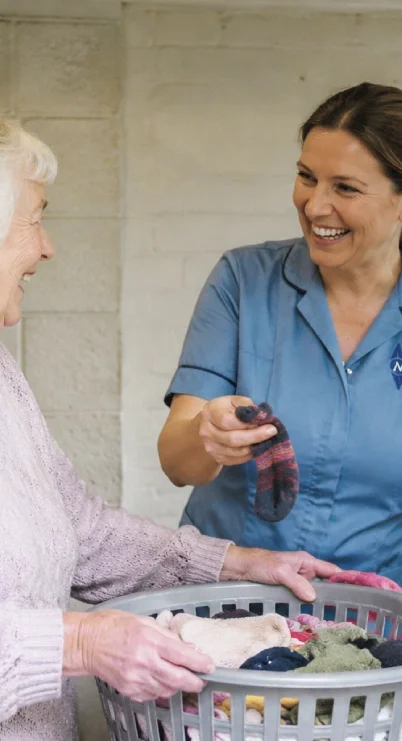 A young woman in a blue shirt laughs while handing a sock to an elderly woman with gray hair, who is holding a laundry basket filled with clothes.