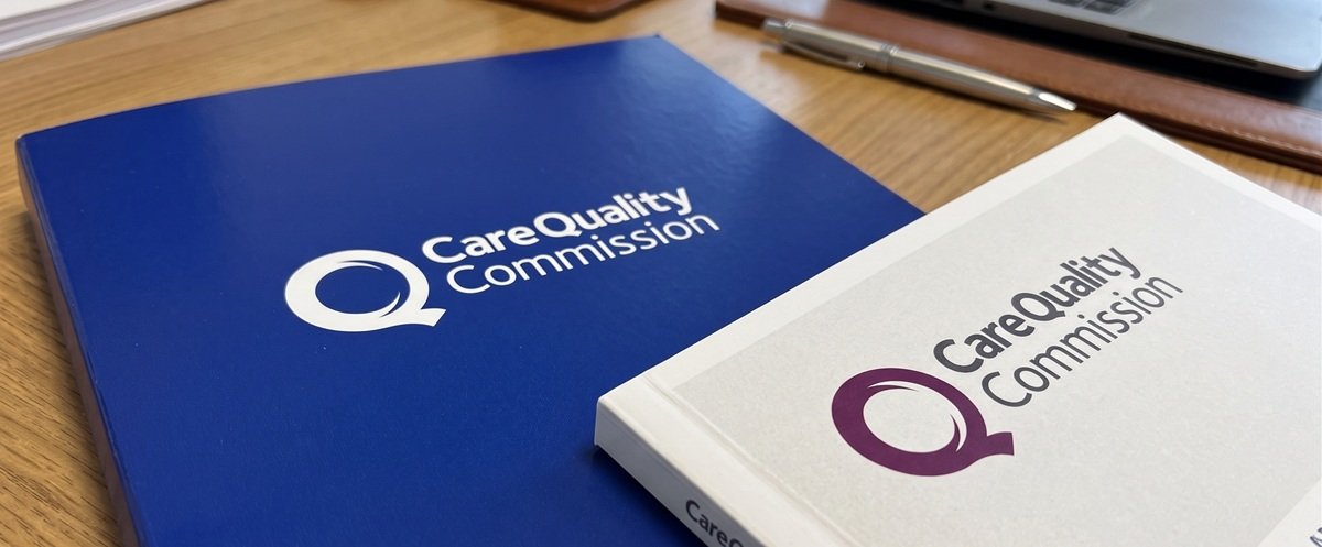 Blue and white folders or binders with the logo and name of the Care Quality Commission on a wooden desk.
