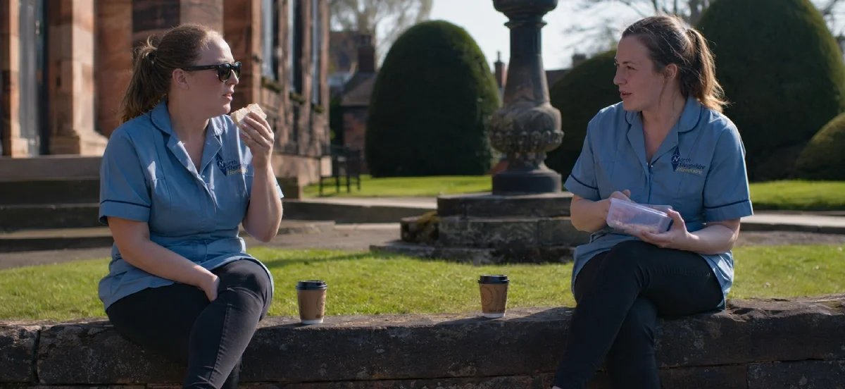 Two women in blue uniforms sitting outside on a stone ledge, having a conversation with coffee cups nearby, in a park with trimmed bushes and historic buildings in the background.