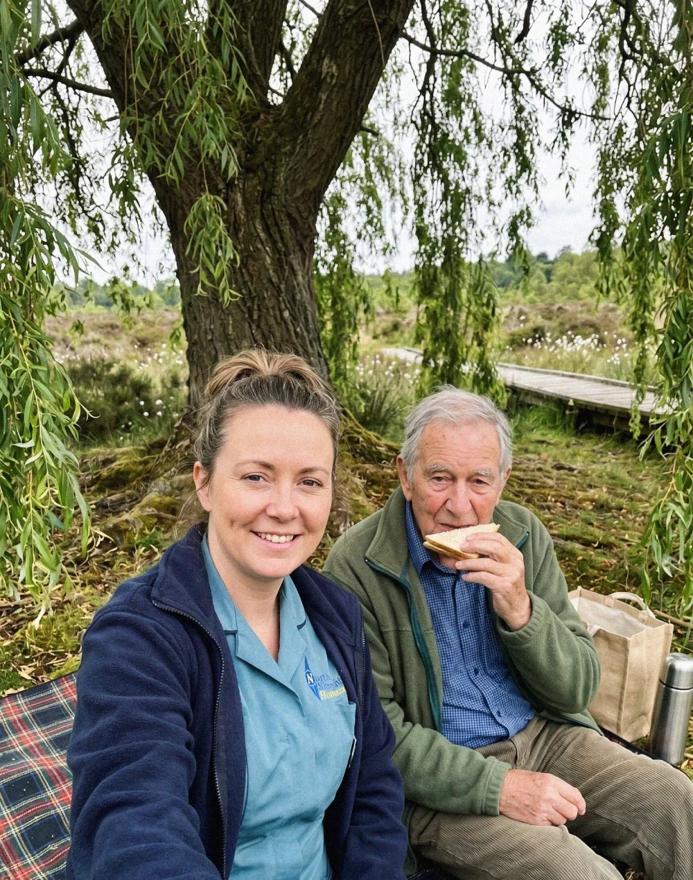 A woman and an older man sitting under a large tree outdoors, enjoying a picnic. The woman is smiling at the camera, while the man is eating a sandwich. There is a blanket on the ground and a bag nearby.