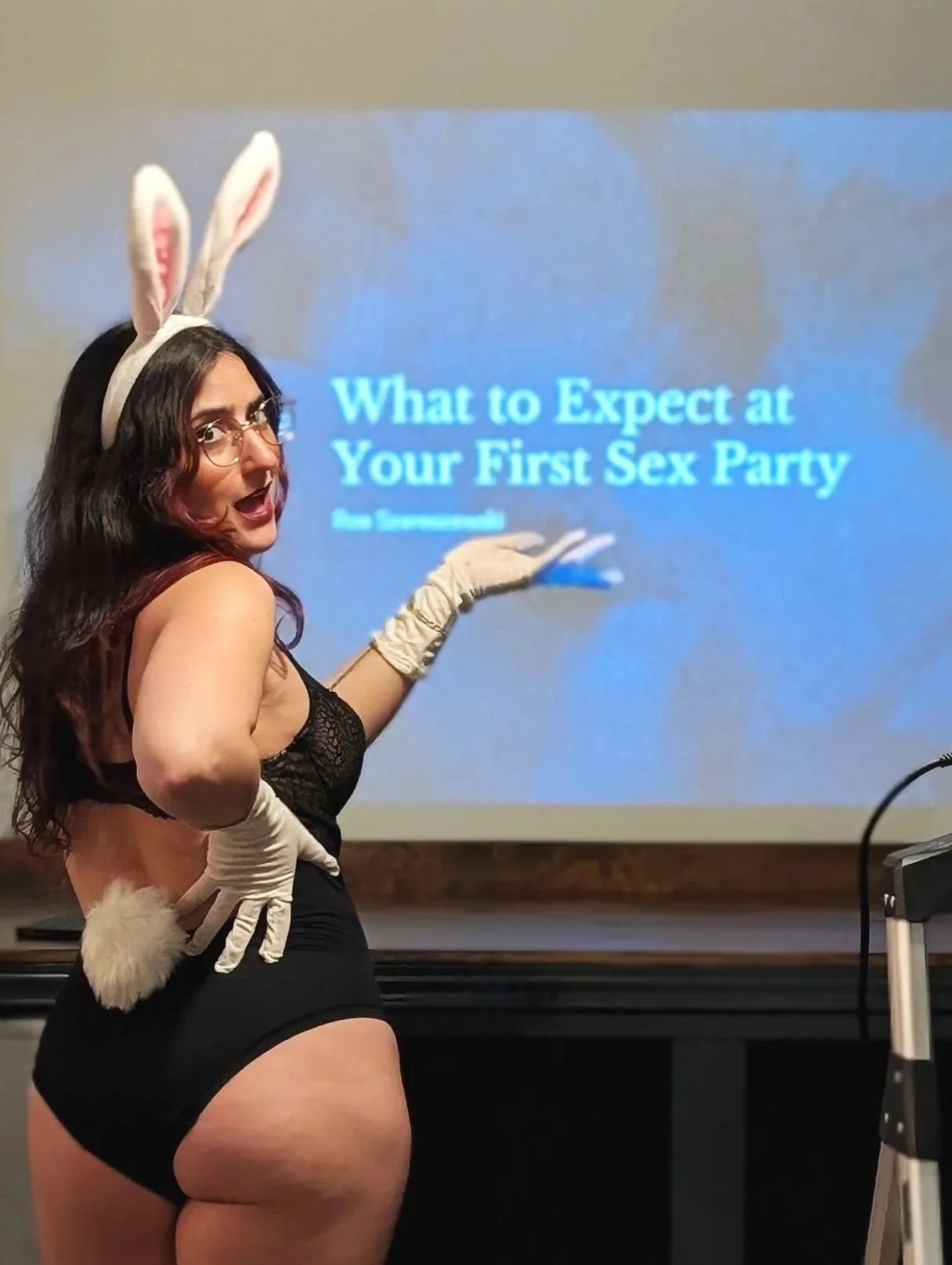 Rae stands in a bunny costume in front of a projector that says, "What to expect at your first sex party"