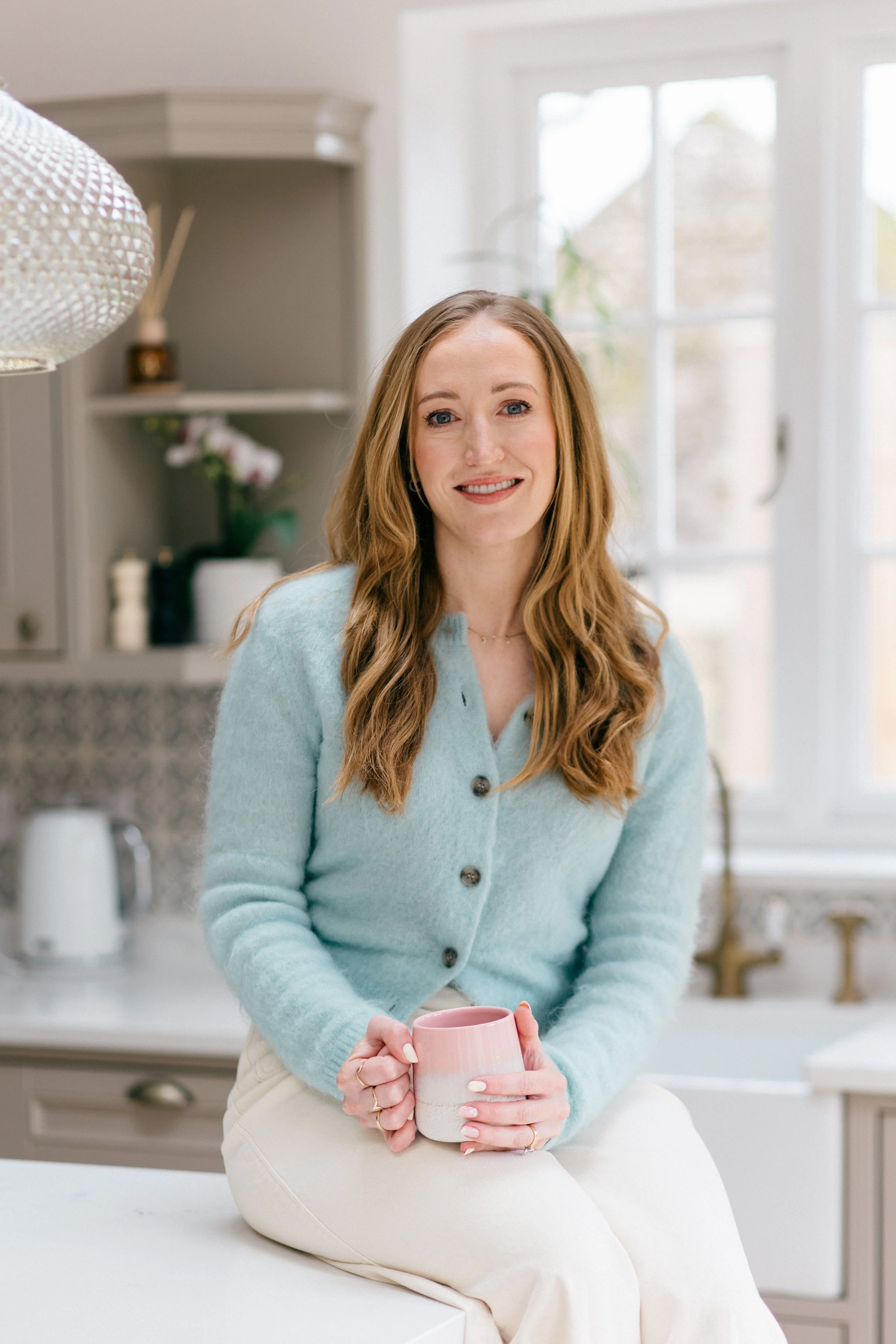 A woman with long red hair and wearing a light blue cardigan, sitting in a bright kitchen, holding a pink and white mug.