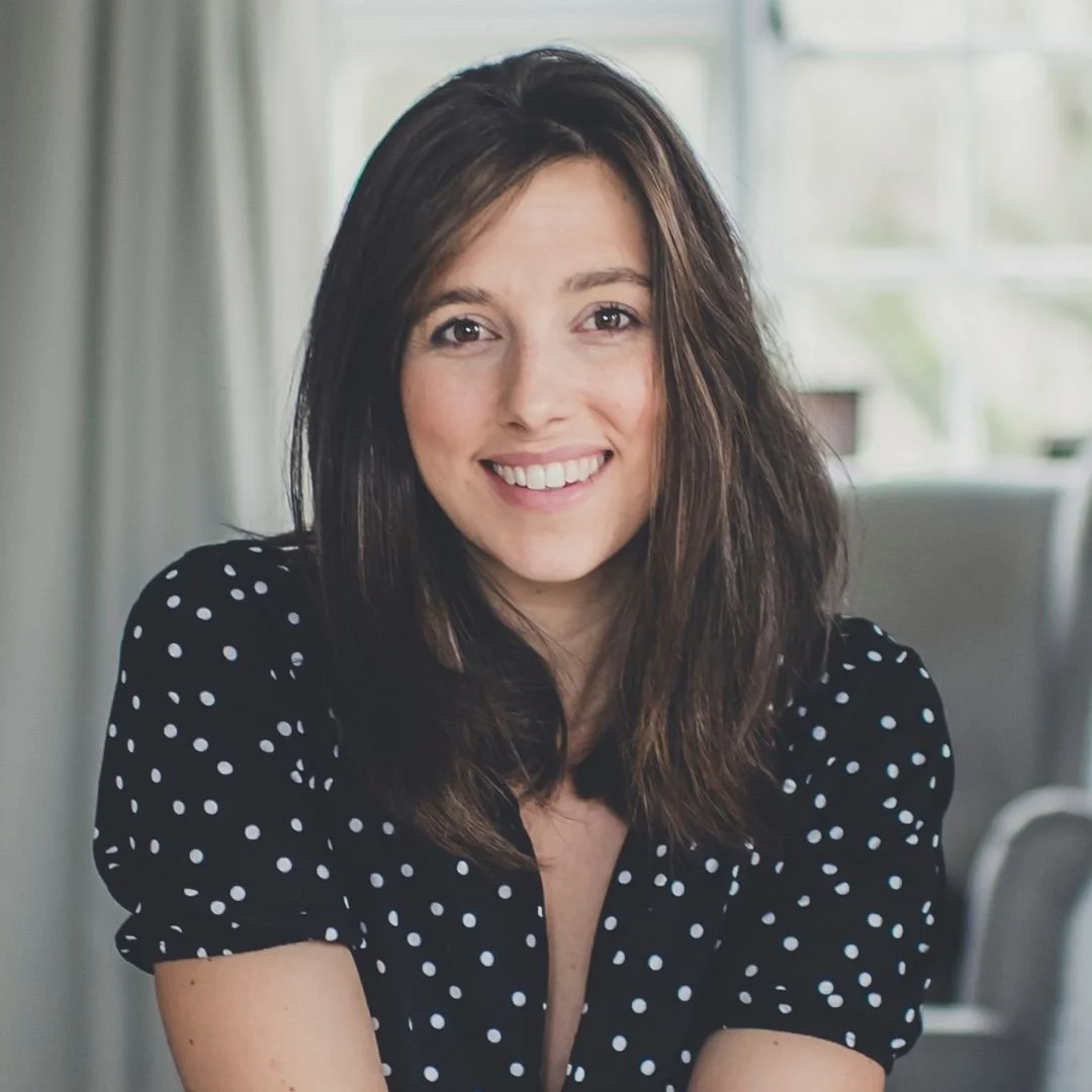 A woman with medium-length brown hair, wearing a black shirt with white polka dots, smiling and sitting indoors with a bright window and light-colored furniture in the background.