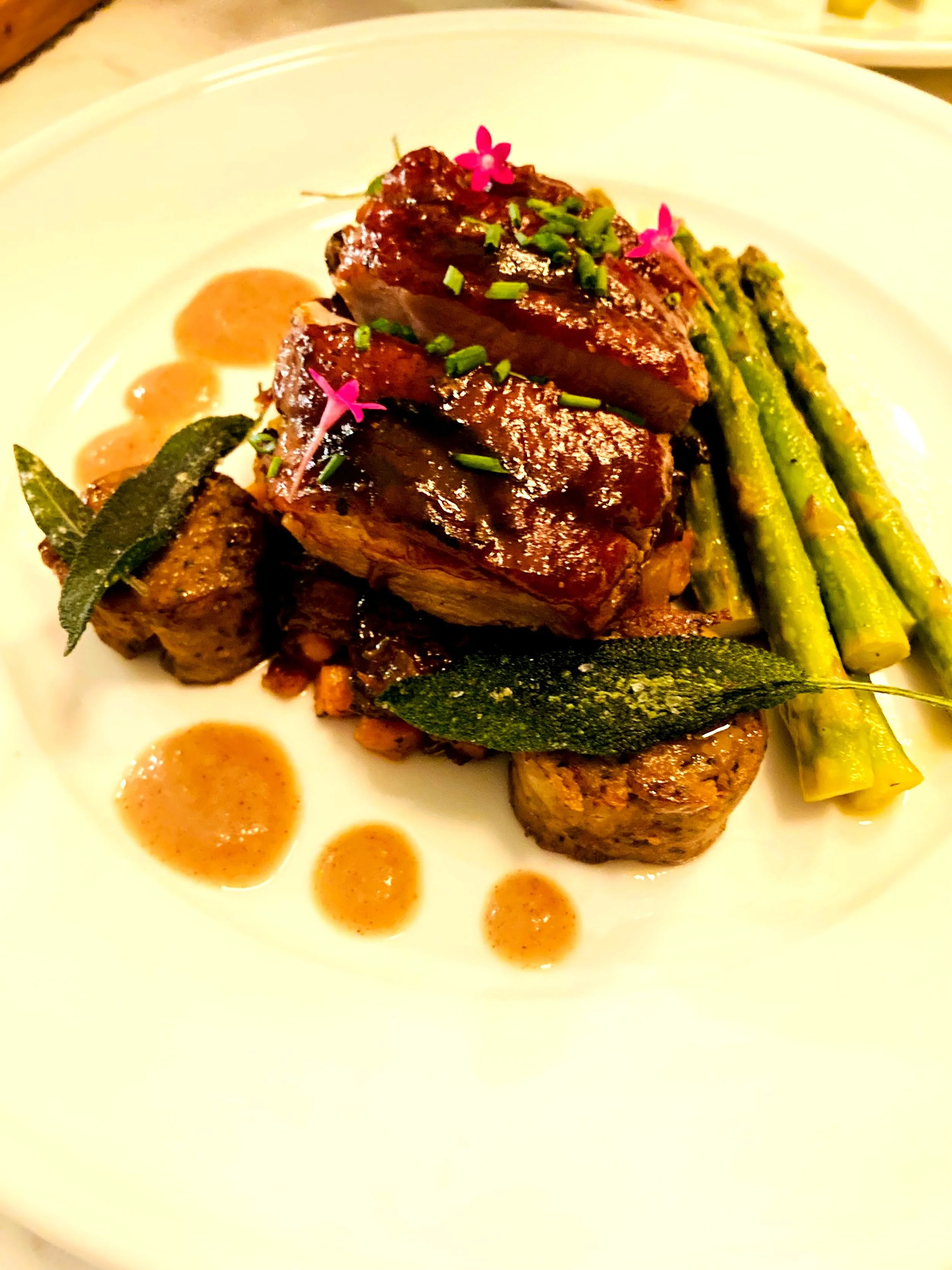 A plated dish with two slices of glazed, roasted meat garnished with green herbs and pink edible flowers, accompanied by sautéed asparagus, bay leaves, and dollops of sauce.