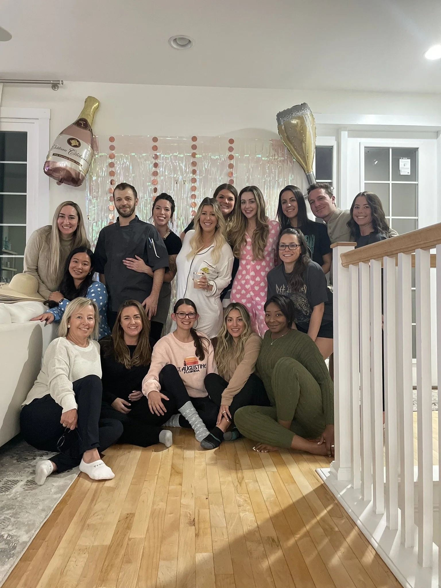 Group of people celebrating a party in a living room, with some dressed in pajamas, balloons, and a decorated backdrop.