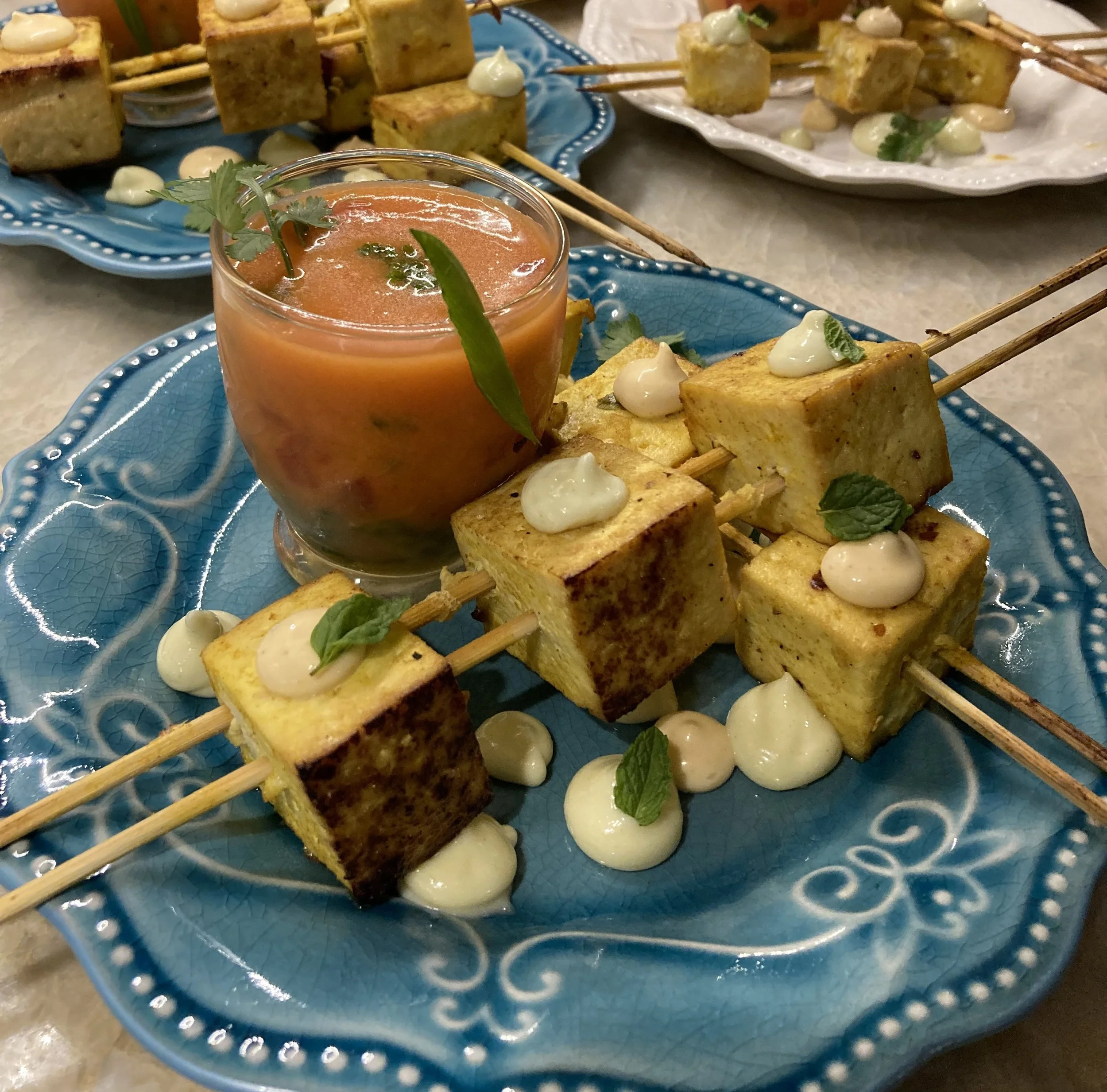 Skewered tofu cubes with sauce and a glass of tomato-based beverage on a decorative blue plate.