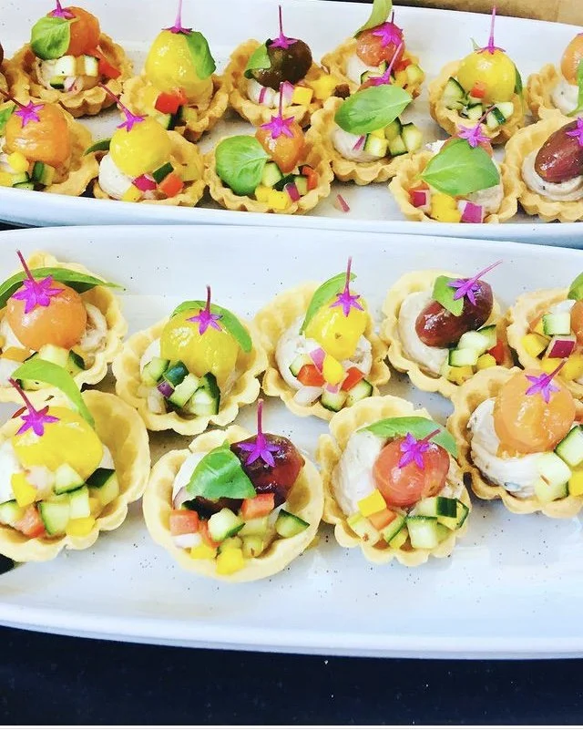 Small appetizer tarts topped with diced vegetables, cubes of cheese, small colorful flowers, and basil leaves on white long plates.
