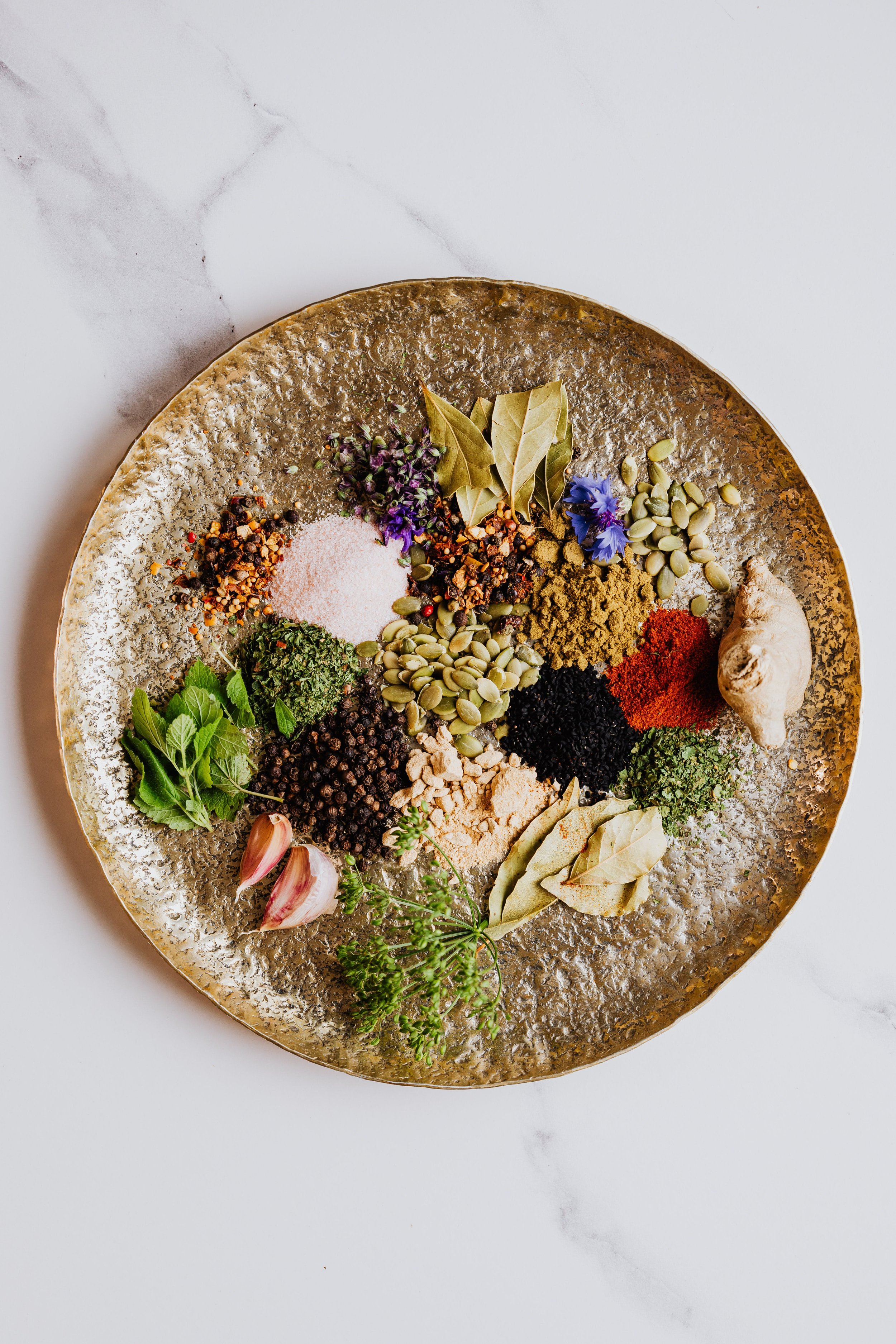 A round, textured gold plate filled with various spices, herbs, and botanicals, including garlic cloves, ginger root, bay leaves, black pepper, turmeric, paprika, and dried herbs.