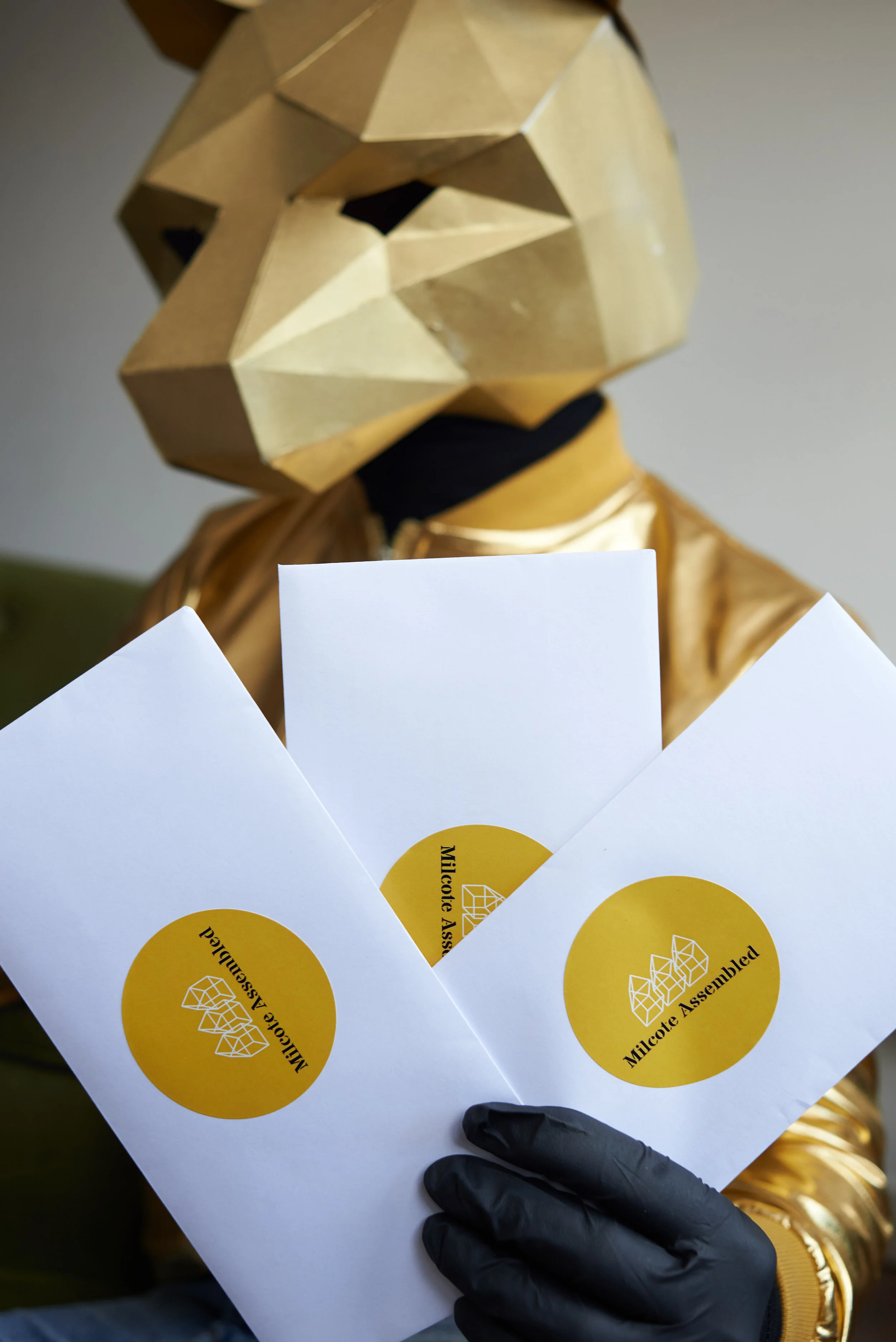 Close-up of someone holding sheets of paper with yellow circles that read 'Milktec Assembled', with a golden, geometrically faceted mask or headgear in the background, and the person wearing a black glove and a golden jacket.