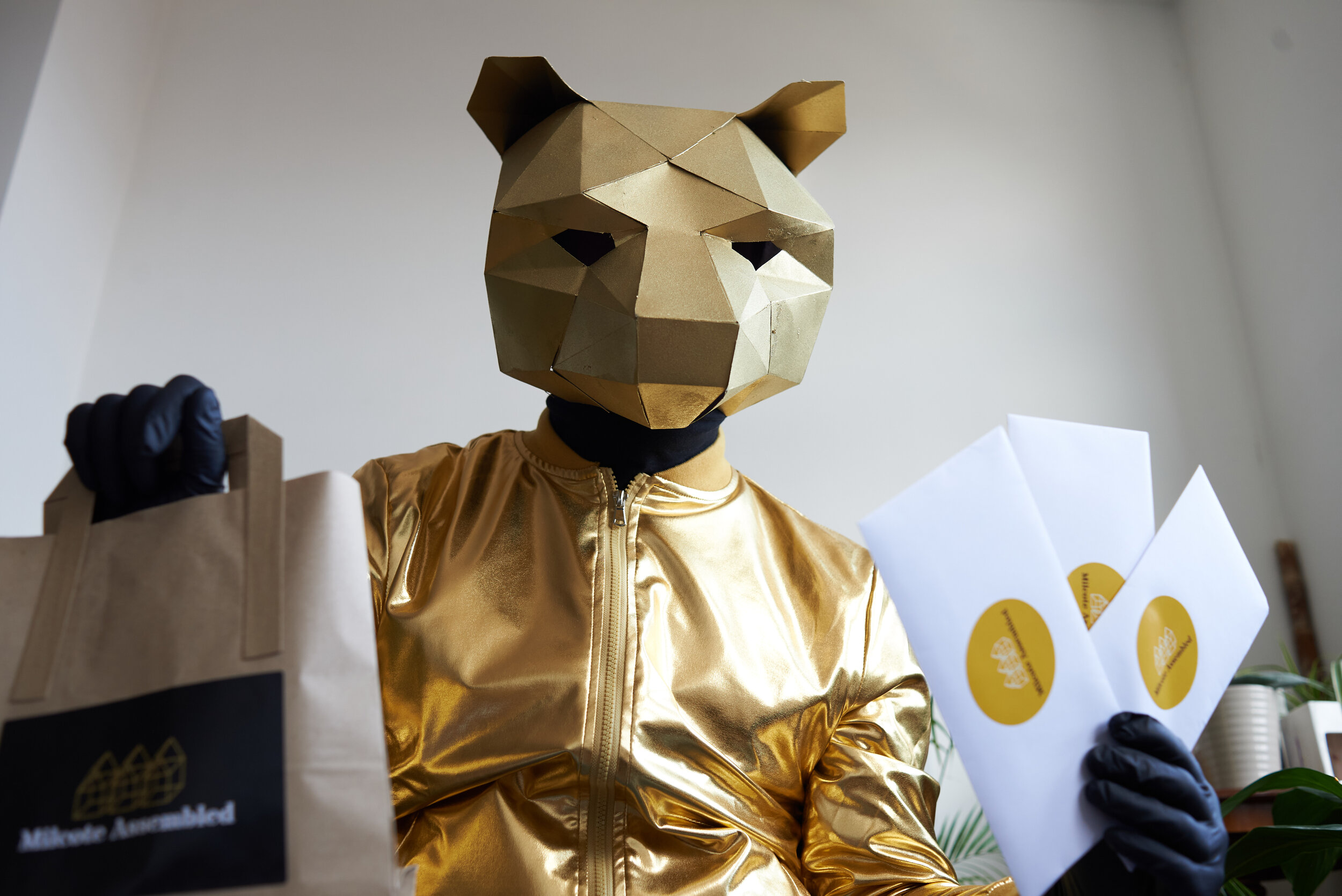 Person wearing a geometric, gold animal mask, gold jacket, and black gloves, holding shopping bags indoors.