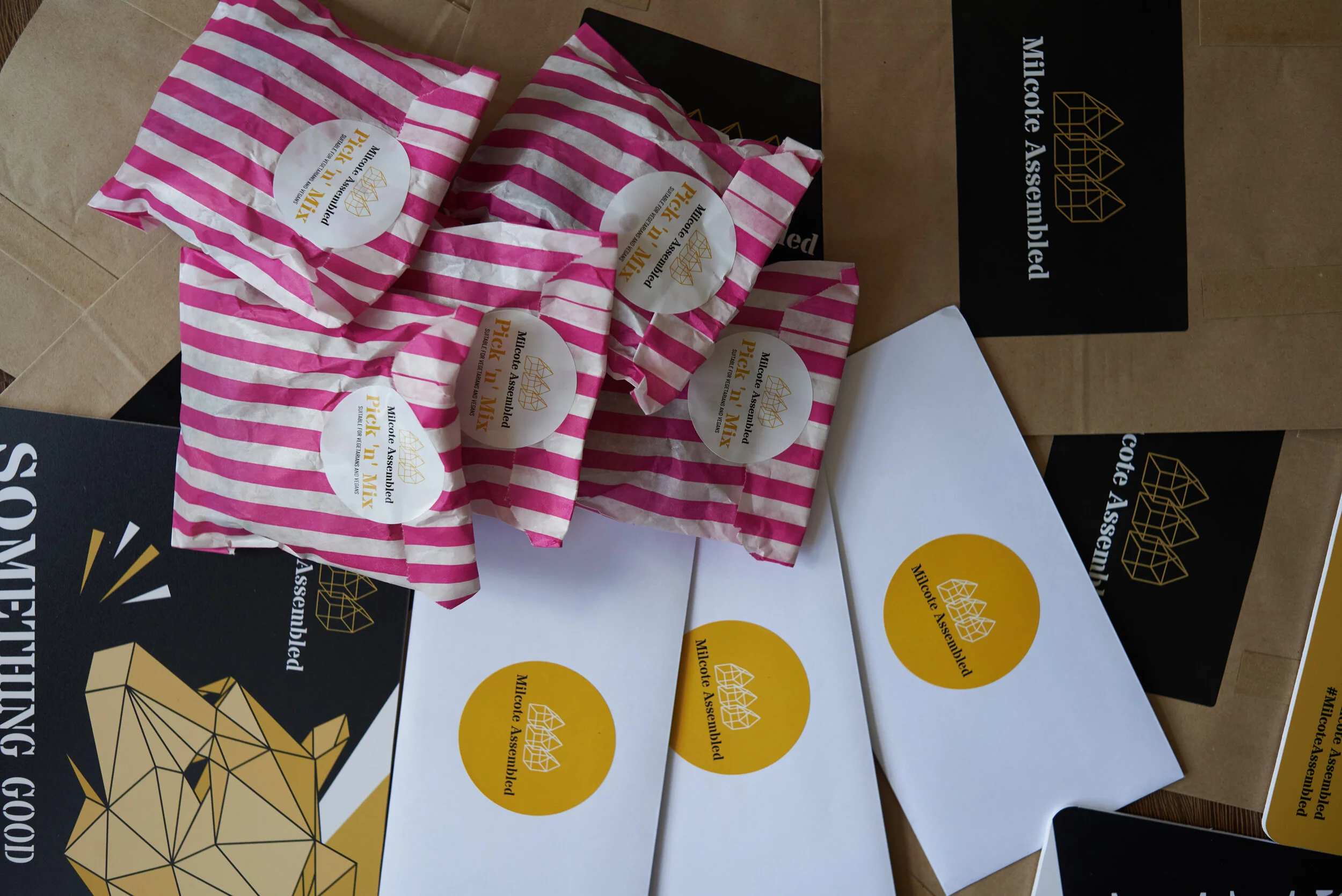 Multiple pink and white striped paper bags with logos, and black and yellow envelopes with logos from a product or brand named Milocote Assembly. The items are arranged on a table.