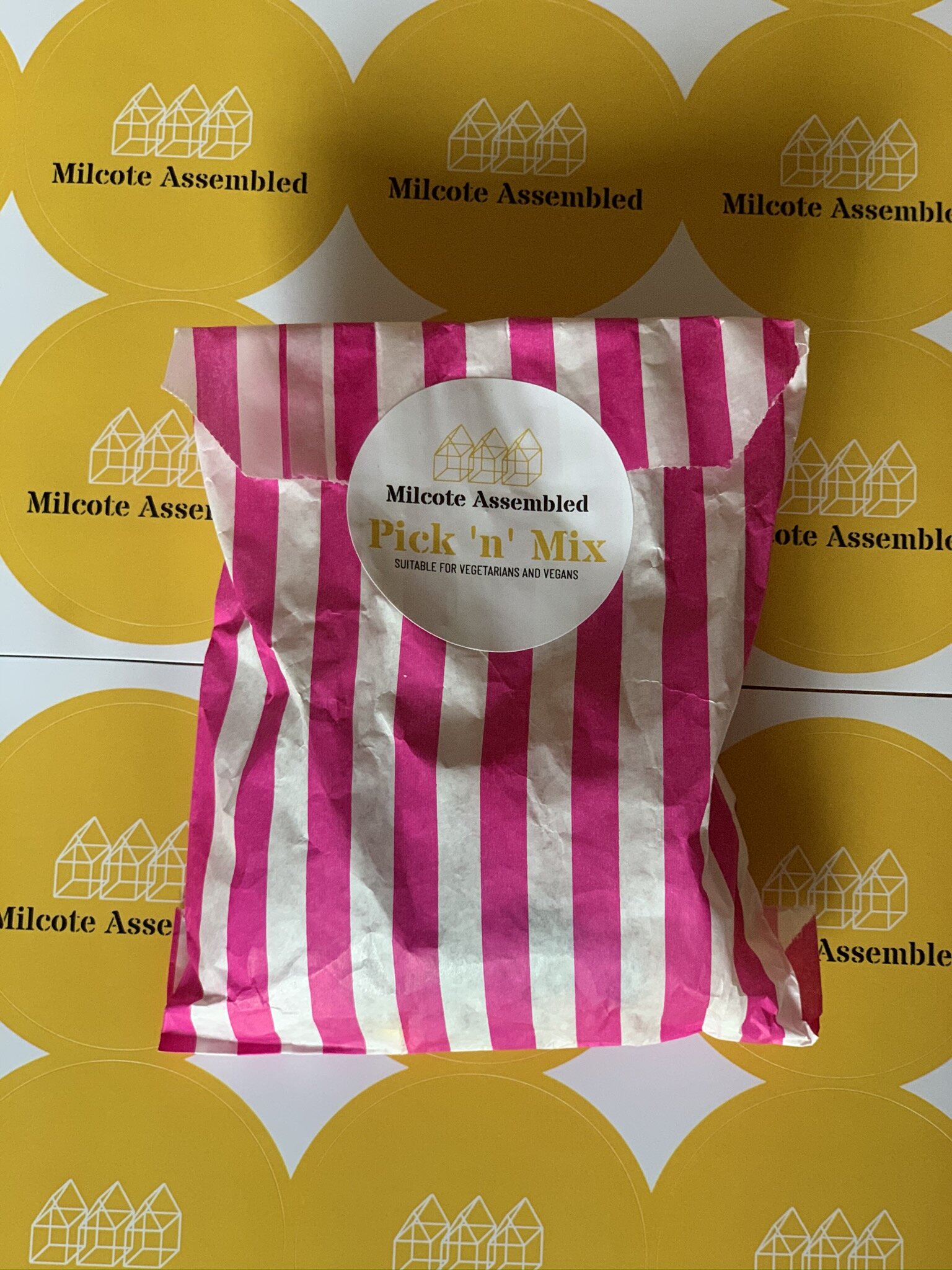 A pink and white striped paper bag labeled 'Pick 'n' Mix' from Milcote Assemblied, placed on yellow circular stickers with the Milcote Assemblied logo.